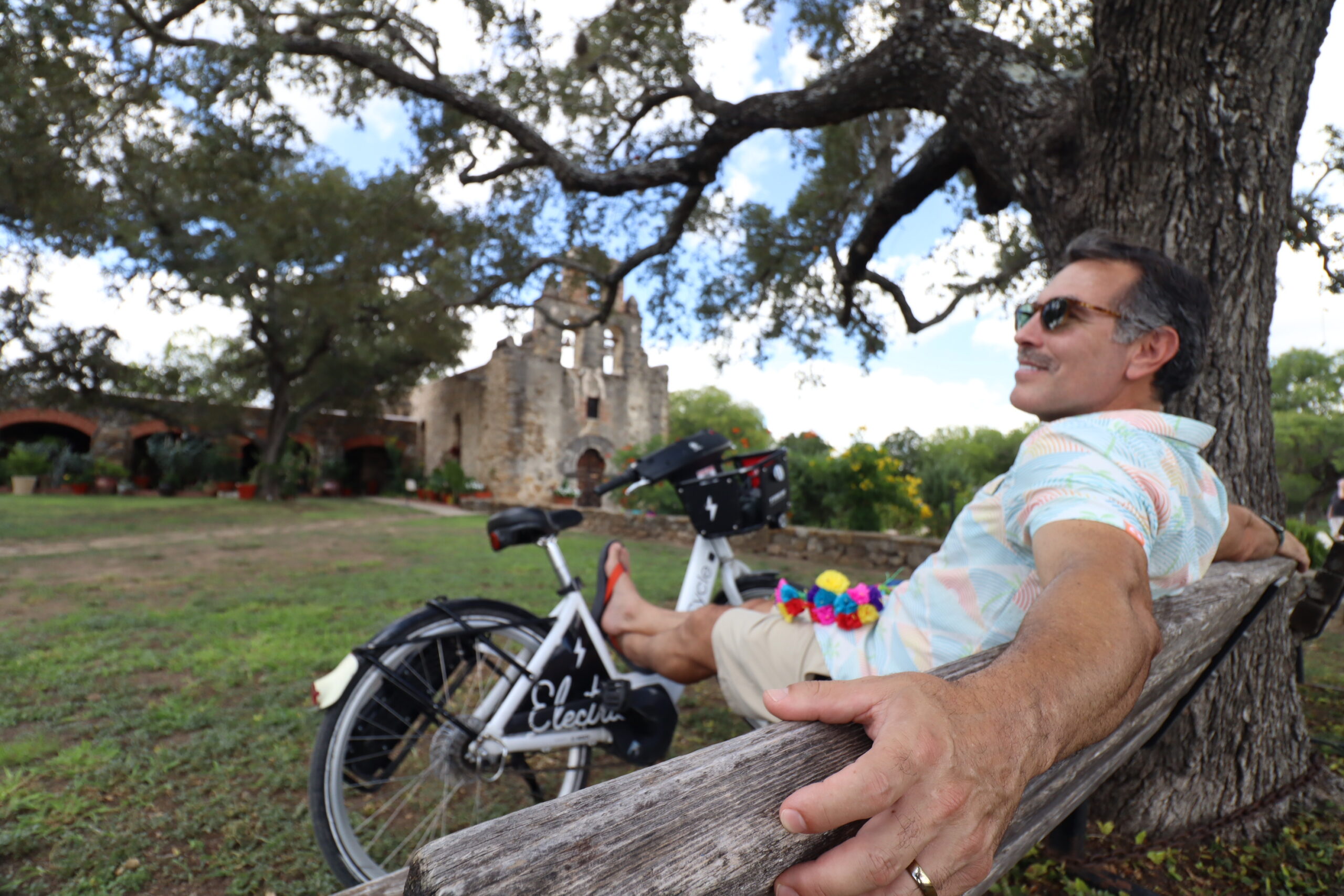 Latino man resting on a bench in front of Mission Espada with a bike share bicycle